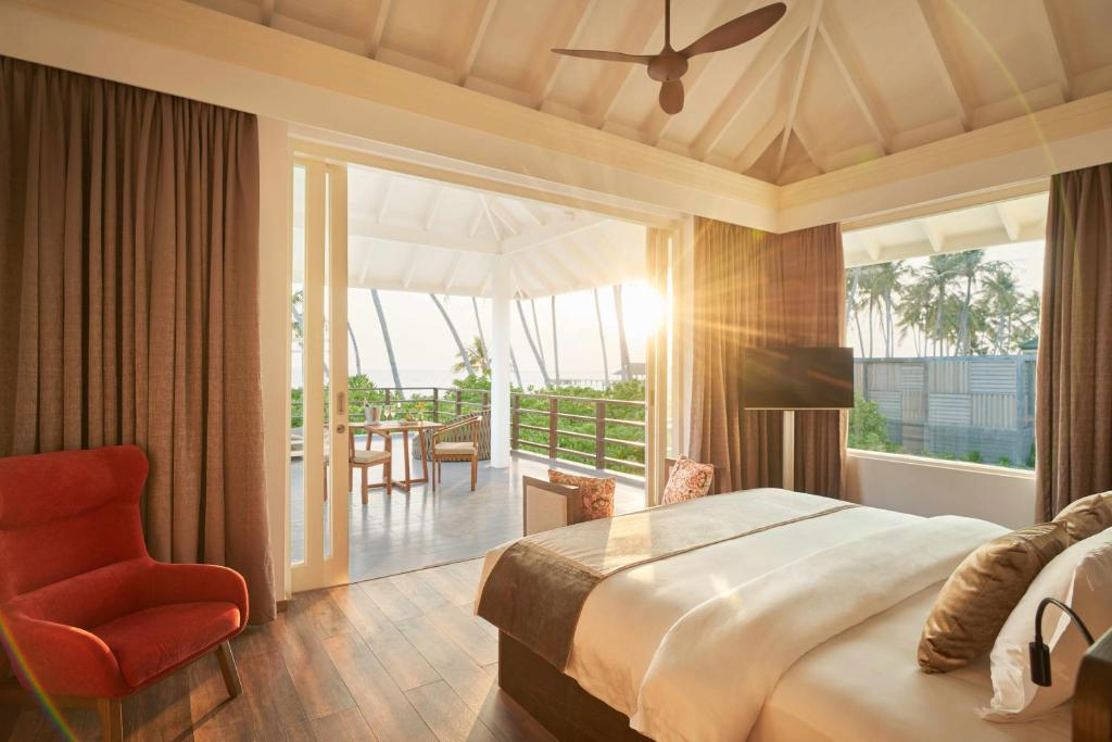 Siyam World Maldives villa interior with king bed, red armchair and doors opening onto a private overwater deck at sunset