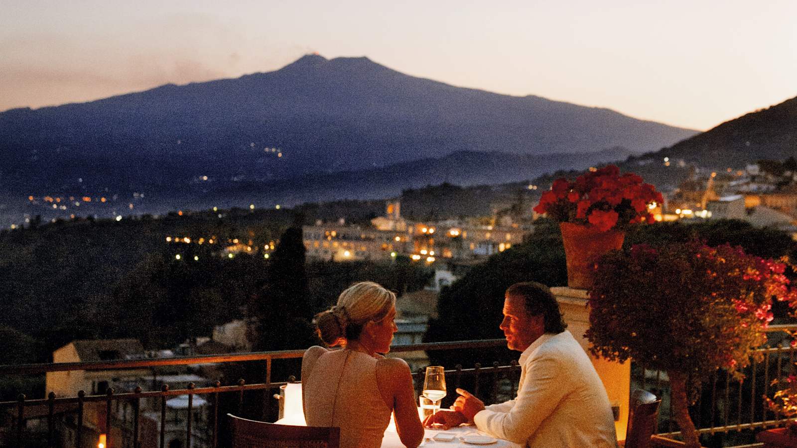 Couple dining by candlelight on the terrace of Grand Hotel Timeo at dusk, with Taormina's lights below and the silhouette of Mount Etna behind