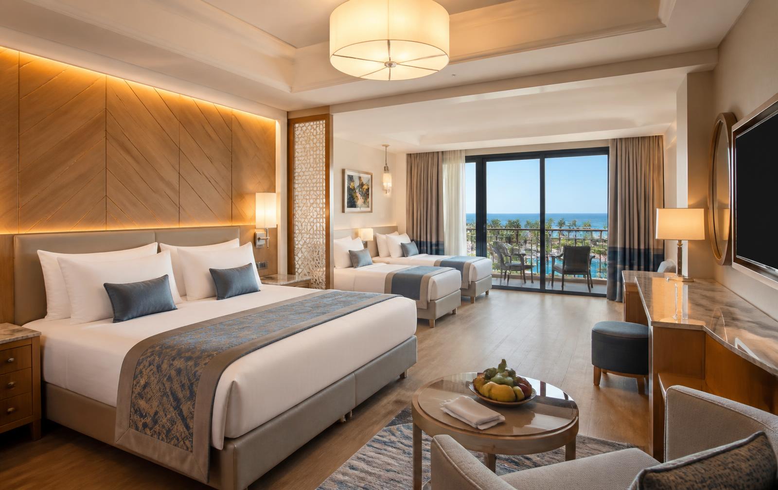 Family suite at a five-star Antalya resort hotel with king bed, twin beds, wood and cream interiors and a balcony overlooking pools and the sea