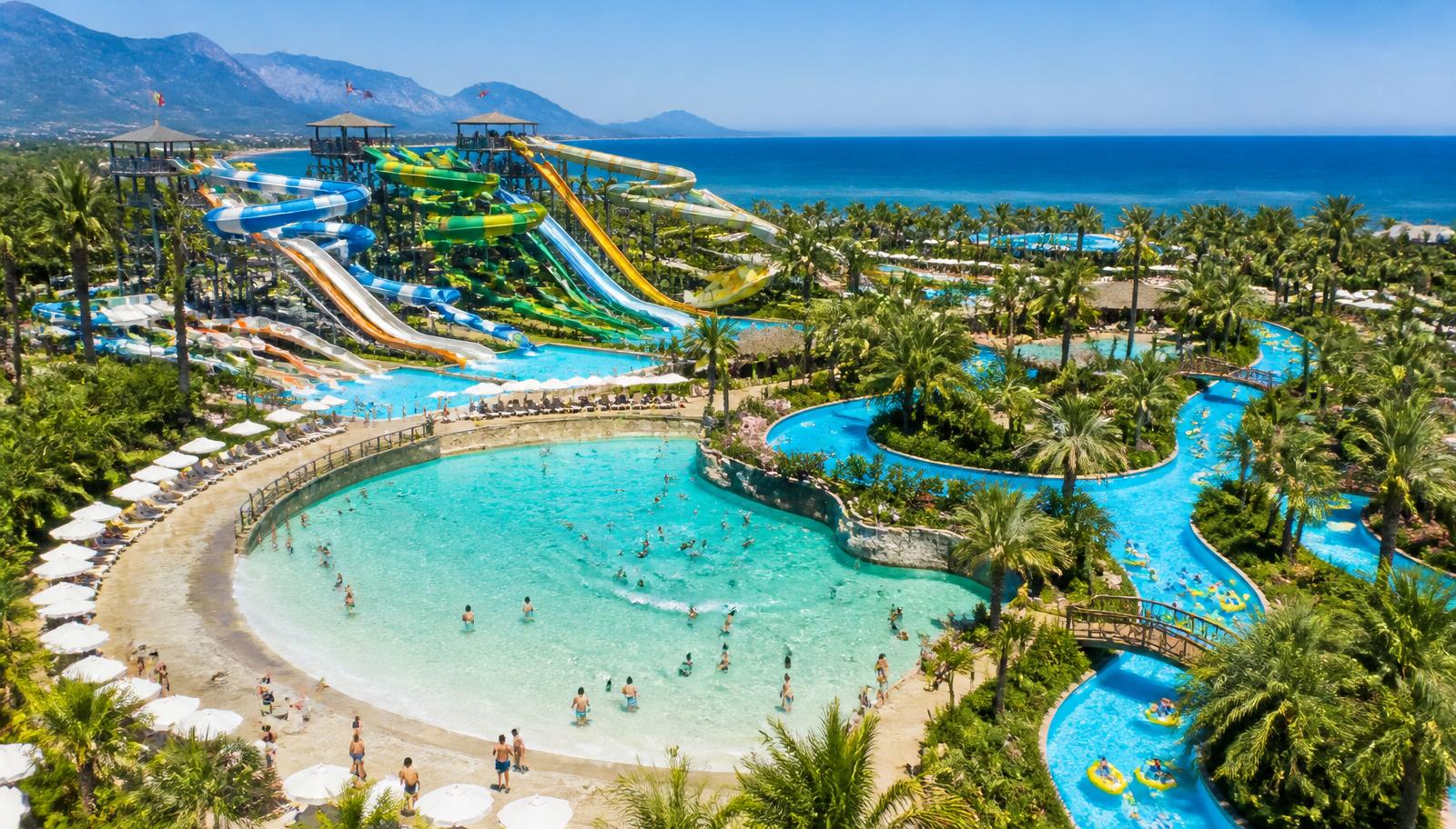 Aerial view of a large aquapark in Antalya with tall twisting waterslides, wave pool, lazy river and palm trees beside the Mediterranean