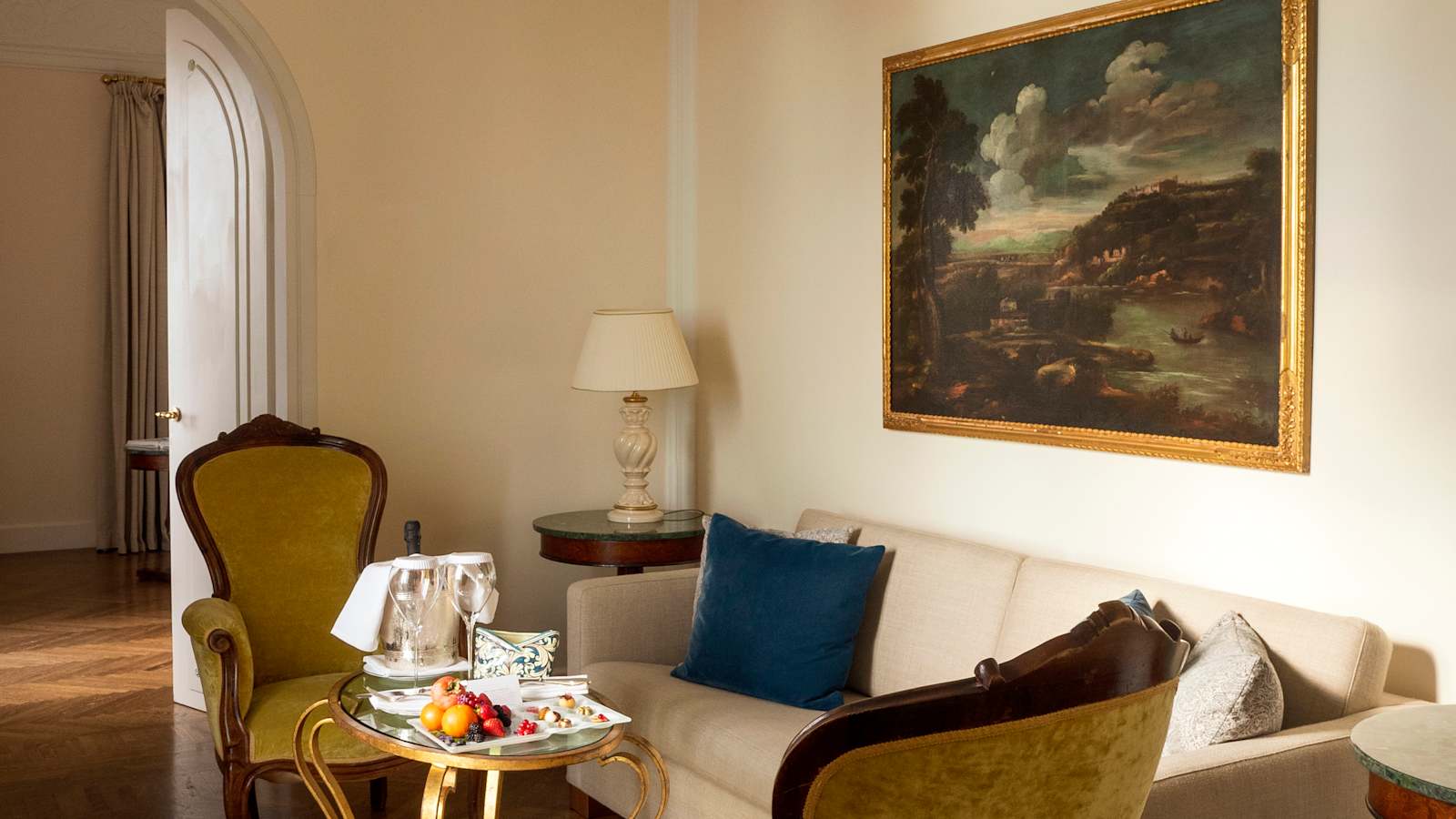 Suite sitting room at Belmond Grand Hotel Timeo with antique gilt-framed landscape painting, velvet armchairs and a welcome fruit and champagne tray