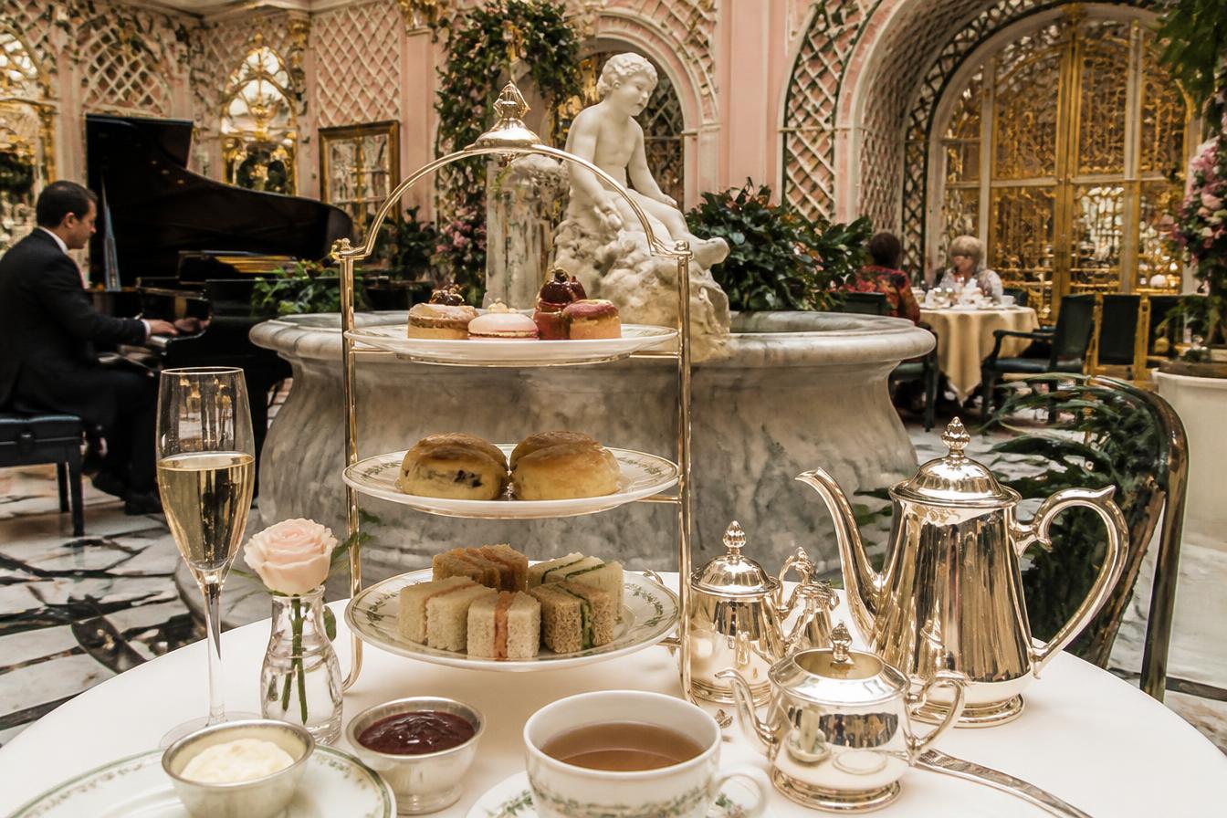 Afternoon tea with tiered cake stand, scones and silver teapot in a pink and gold London hotel palm court with pianist